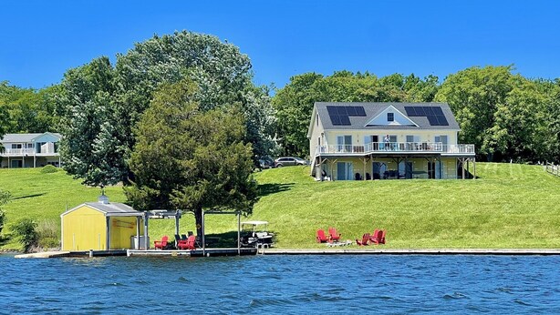 Una propiedad junto al lago con una casa grande, un muelle con sillas y un cobertizo pequeño.