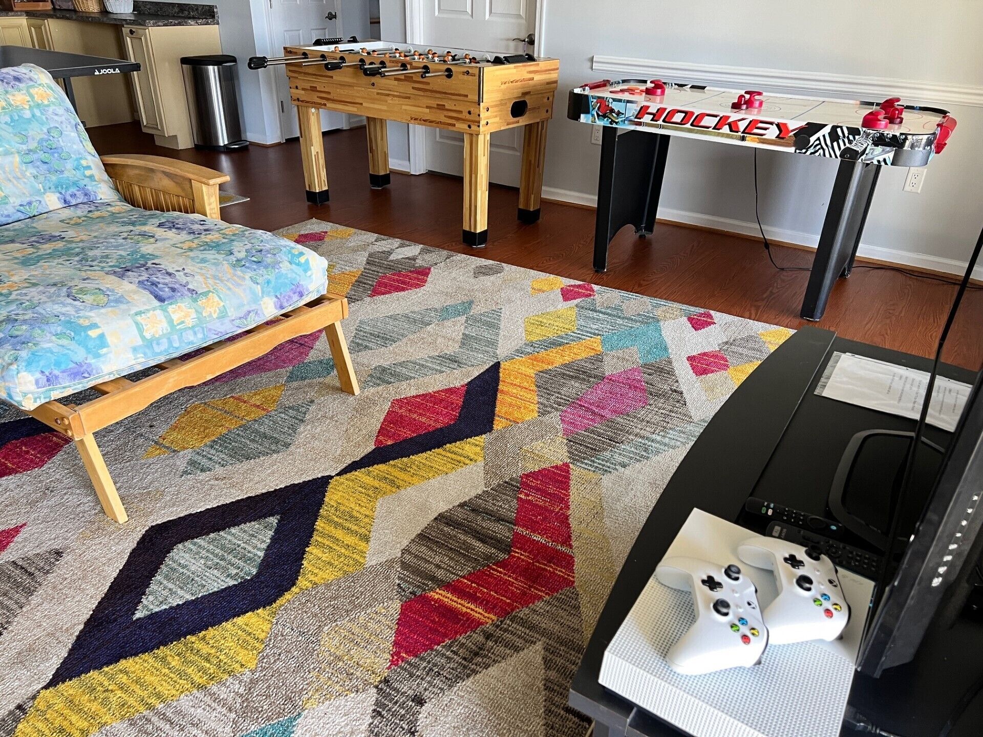 Ein gemütliches Wohnzimmer mit einem farbenfrohen Teppich, einem Holztisch, einer Couch, einem Kicker und einer Spielekonsole mit Controllern.