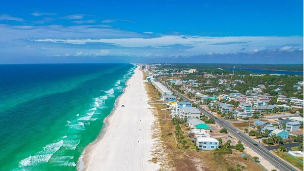 Vista aérea de un pueblo costero con una extensa playa arenosa, una carretera que sigue la línea de la costa y edificios residenciales de diversos estilos arquitectónicos.