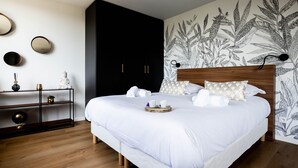 Un lit bien fait, avec du linge de lit blanc, et un plateau contenant des articles pour le petit-déjeuner. On y trouve une armoire sombre, une tête de lit en bois et un mur orné d’une fresque représentant des motifs végétaux.