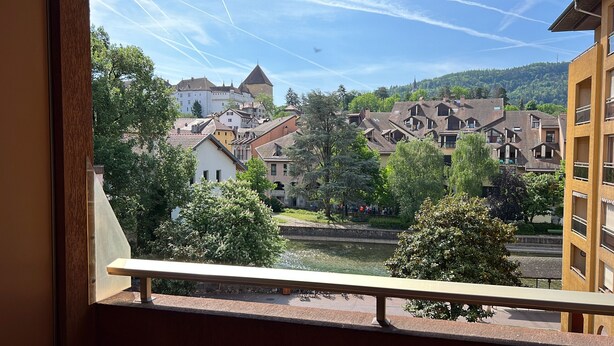 Une vue depuis un balcon donnant sur une rivière, avec des bâtiments, dont une structure ressemblant à un château, et des arbres.