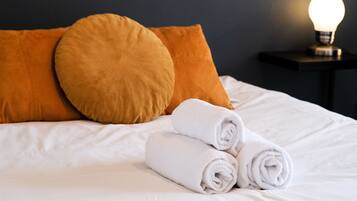 Ein ordentlich bezogenes Bett mit weißen Leinentüchern, zwei aufgerollten Handtüchern und zwei orangenen Kissen.