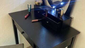 Une petite coiffeuse de couleur sombre, dotée d’un miroir et de lampes, ainsi qu’une tasse et des produits de maquillage.