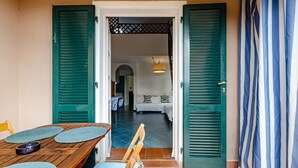 Quarto com uma mesa de madeira, duas portas verdes, uma cama com almofadas e chão de azulejos.