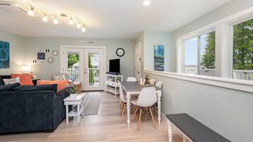 A modern living room with a dark sofa, white chairs, and a dining table. There is a bench along the window, a clock on the wall, and a sign that reads "CRYSTALLAKE CALM".