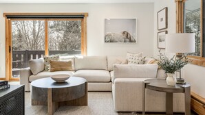 A living room with a sofa, a coffee table, and a side table. There is a window with a view of trees and a framed picture on the wall. - Luxury Vail 3bd/2ba w/ Hot Tub! (Vail)