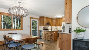 A modern kitchen with wooden cabinets, a central island with stools, a dining area with a wooden table and chairs, and a large window with a view of trees. - Luxury Vail 3bd/2ba w/ Hot Tub! (Vail)