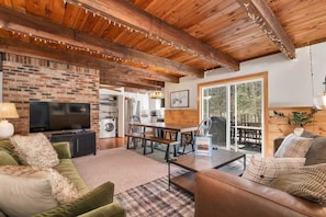 A cozy living room with a brick fireplace, wooden ceiling beams, a television, and a dining area with a table and chairs.