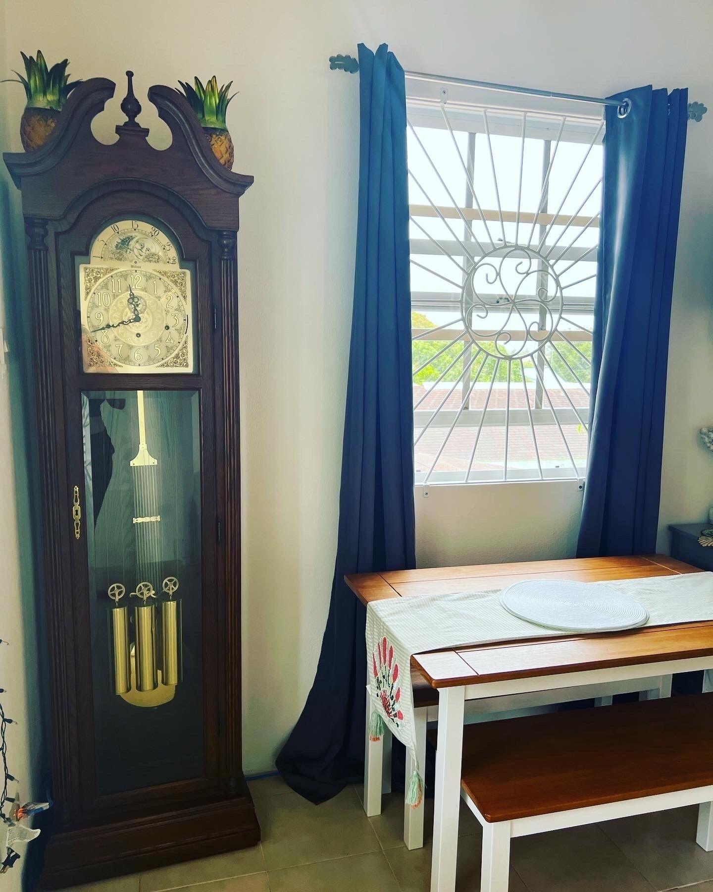 Een kamer met een houten staande klok, een tafel met een bankje, en een raam met blauwe gordijnen.