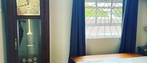 Een kamer met een houten staande klok, een tafel met een bankje, en een raam met blauwe gordijnen.