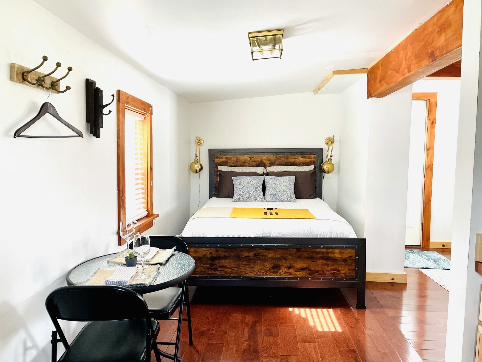 Un dormitorio con cama, lámparas de noche, un perchero, una pequeña mesa con sillas y una ventana con persianas.