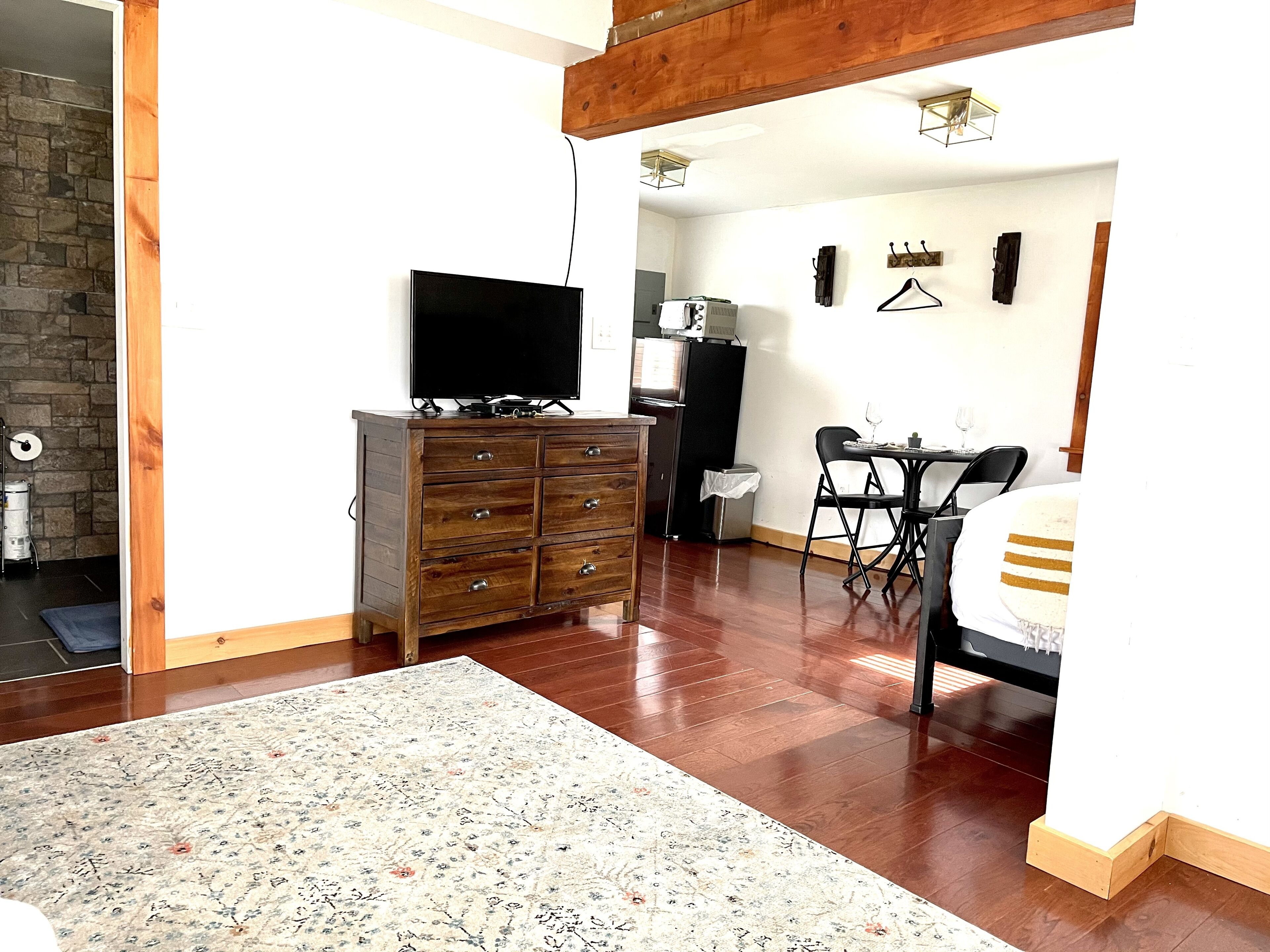 A living room with a wooden dresser, a flat-screen TV, a dining table with chairs, and a kitchen area in the background.