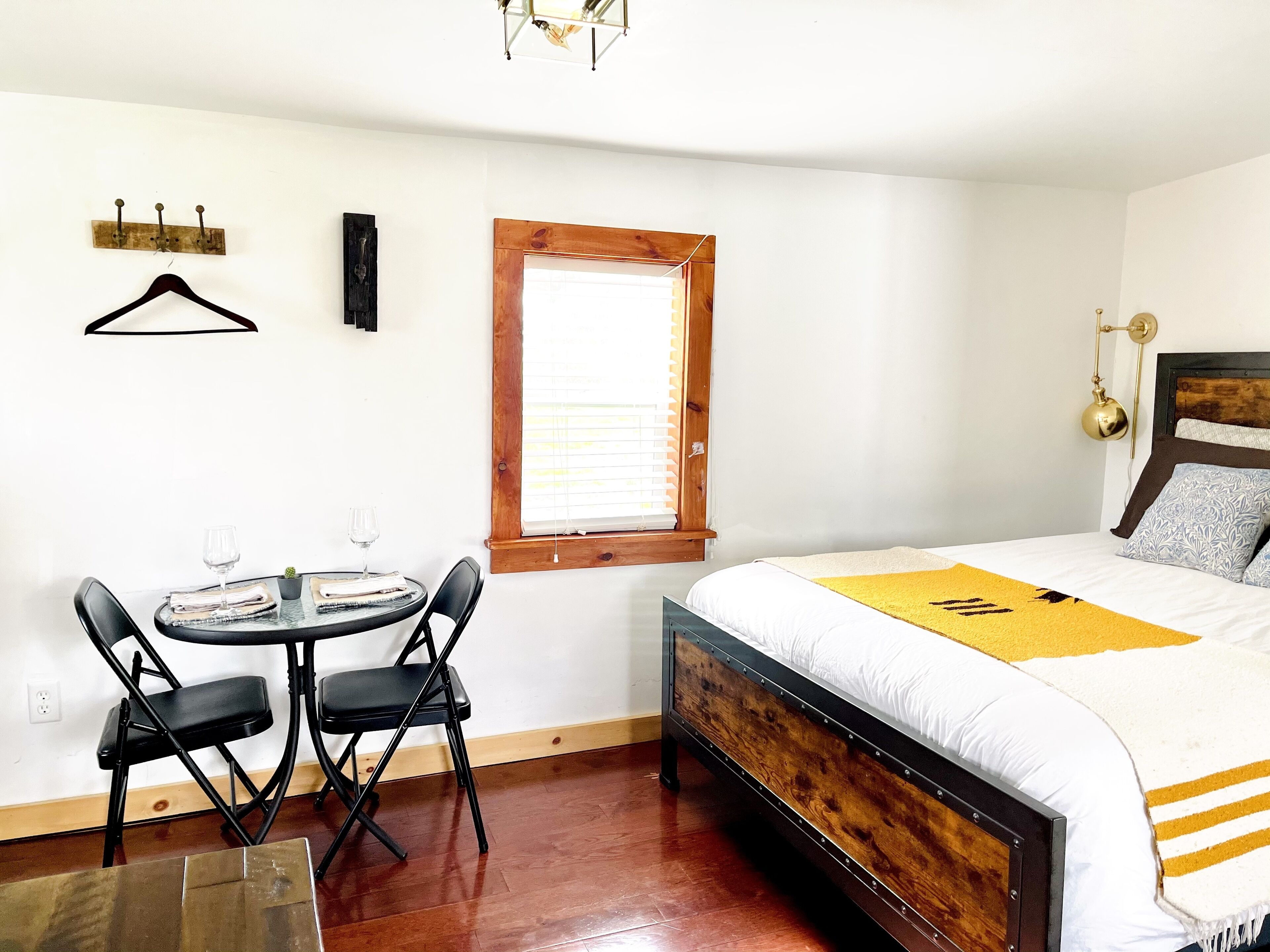 A bedroom with a wooden bed, a small round table with chairs, a window with blinds, and a wall-mounted light fixture.