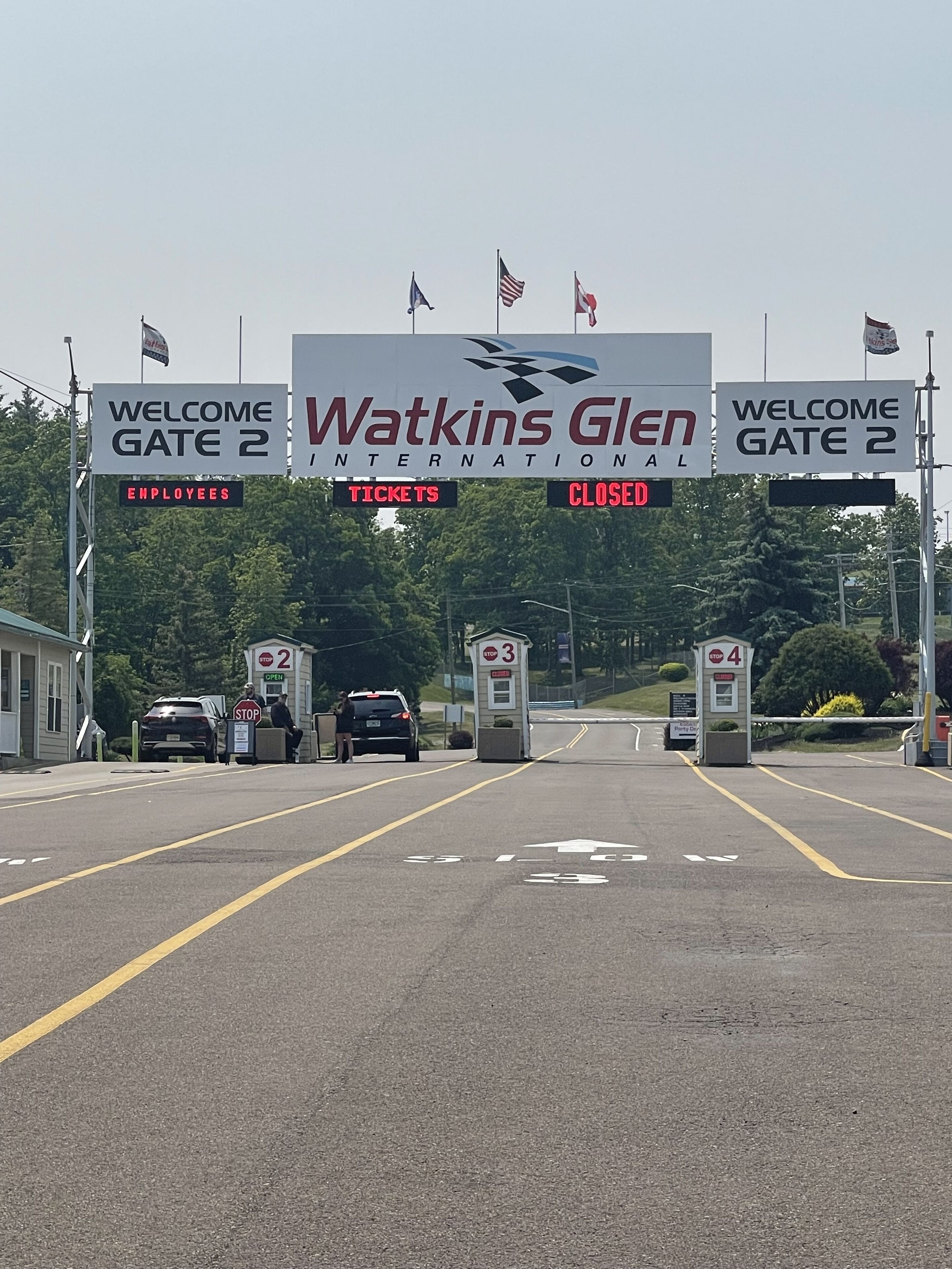 A gate entrance with signs for 'Welcome Gate 2' and 'Watkins Glen International' with flags. There are stop signs and a closed gate.