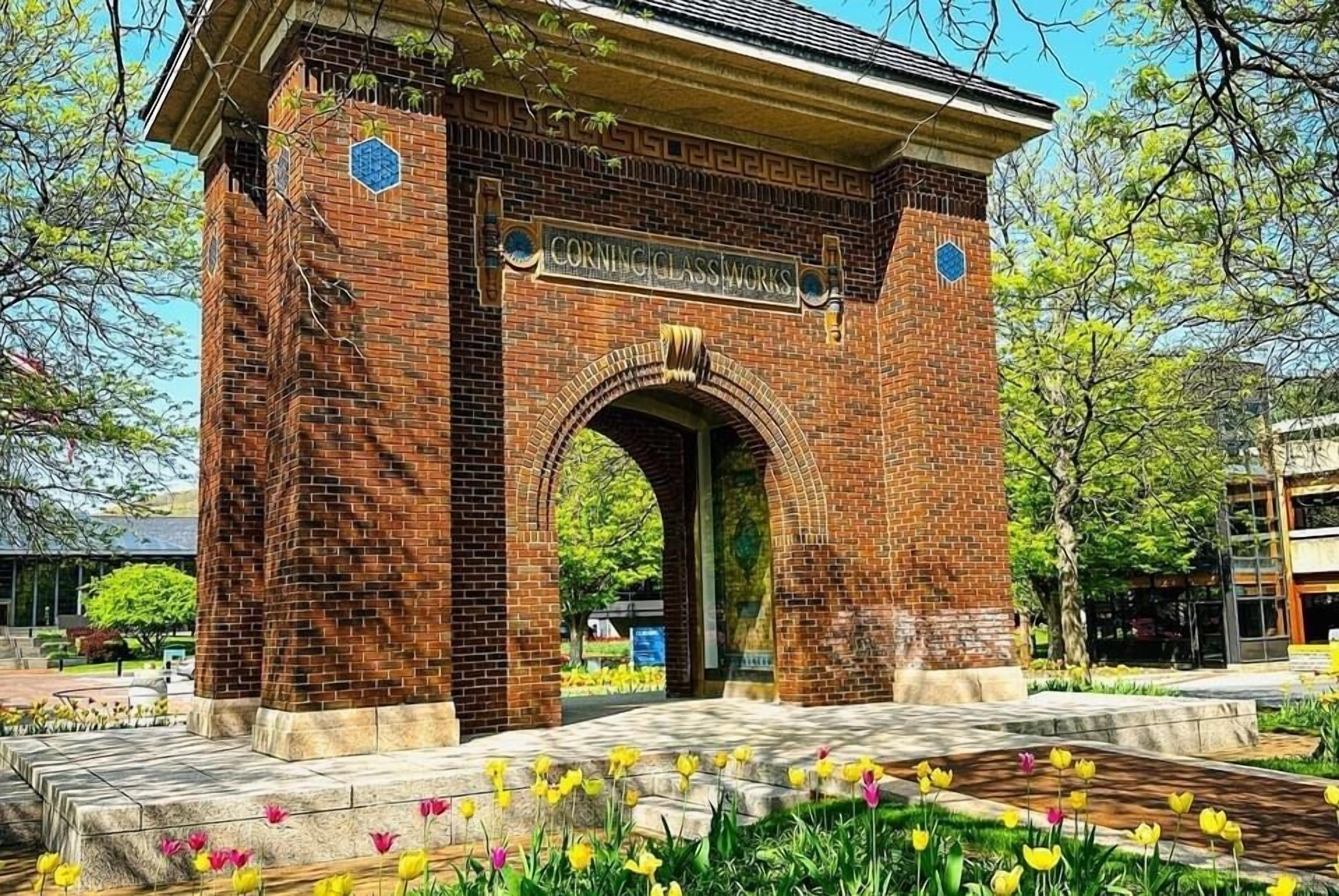 A brick archway with the inscription "Cornick Glass Works" and a surrounding garden with flowers.