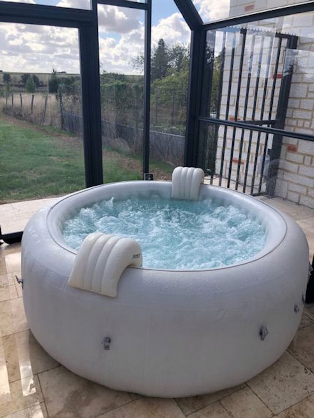 Ein Whirlpool mit zwei integrierten Sitzplätzen, umgeben von einer Glaskonstruktion mit Blick auf eine Rasenfläche und eine Backsteinmauer.