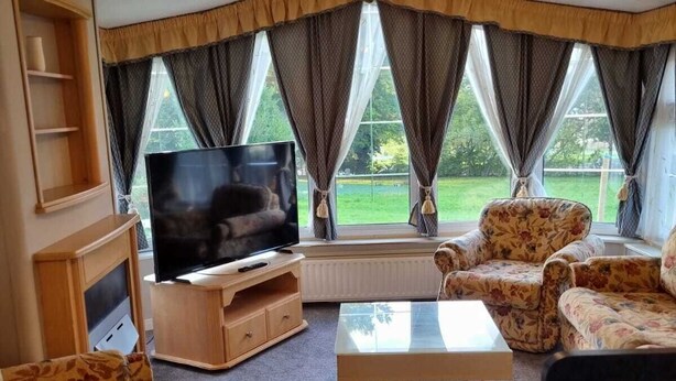 A cozy living room with a flat-screen TV, floral-patterned furniture, and a ceiling fan. - Quite 3 Bedroom Static Caravan (Orpington)