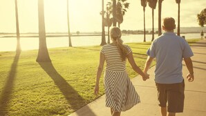 A couple walking hand in hand along a waterfront path with palm trees and a body of water in the background.