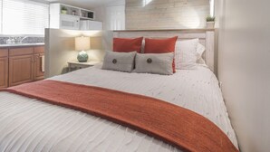 A bedroom with a bed, bedside table, lamp, and a view of the kitchen area.