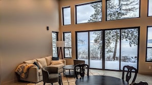 A modern living room with a sofa, chairs, and a dining table. Large windows offer a view of snow-covered trees and a body of water. - Skipping Stone Lake House, Skipping Stone Lake House (Woods Bay)