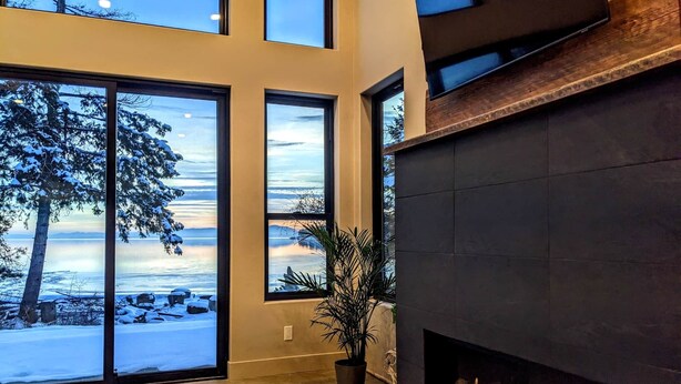 A modern living room with large windows overlooking a snowy landscape, a fireplace, and a potted plant. - Skipping Stone Lake House, Skipping Stone Lake House (Woods Bay)