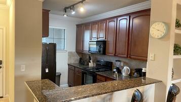 A kitchen with granite countertops, wooden cabinets, a refrigerator, an oven, a microwave, and a clock on the wall.