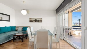 A modern living room with a glass dining table, white chairs, a sofa with teal cushions, and a cityscape painting on the wall. - WELCS APARTAMENTO 218 EMP frente al Mar (Empuriabrava)