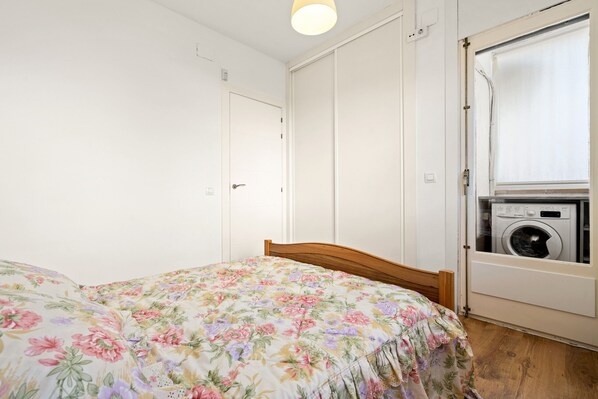Un dormitorio con colcha estampada, cabecera de madera, lavadora en un rincón y una puerta blanca.