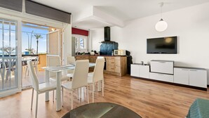 A modern kitchen with a large TV, wooden cabinets, and a dining area with white chairs and a glass table. - WELCS APARTAMENTO 218 EMP frente al Mar (Empuriabrava)
