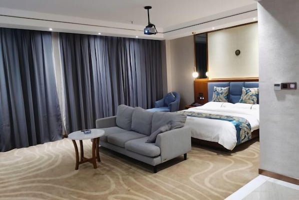 A modern hotel room with a grey sofa, a small round table, a bed with a floral bedspread, and a large window with curtains. - Jingan Haoting Hotel (Gengma)