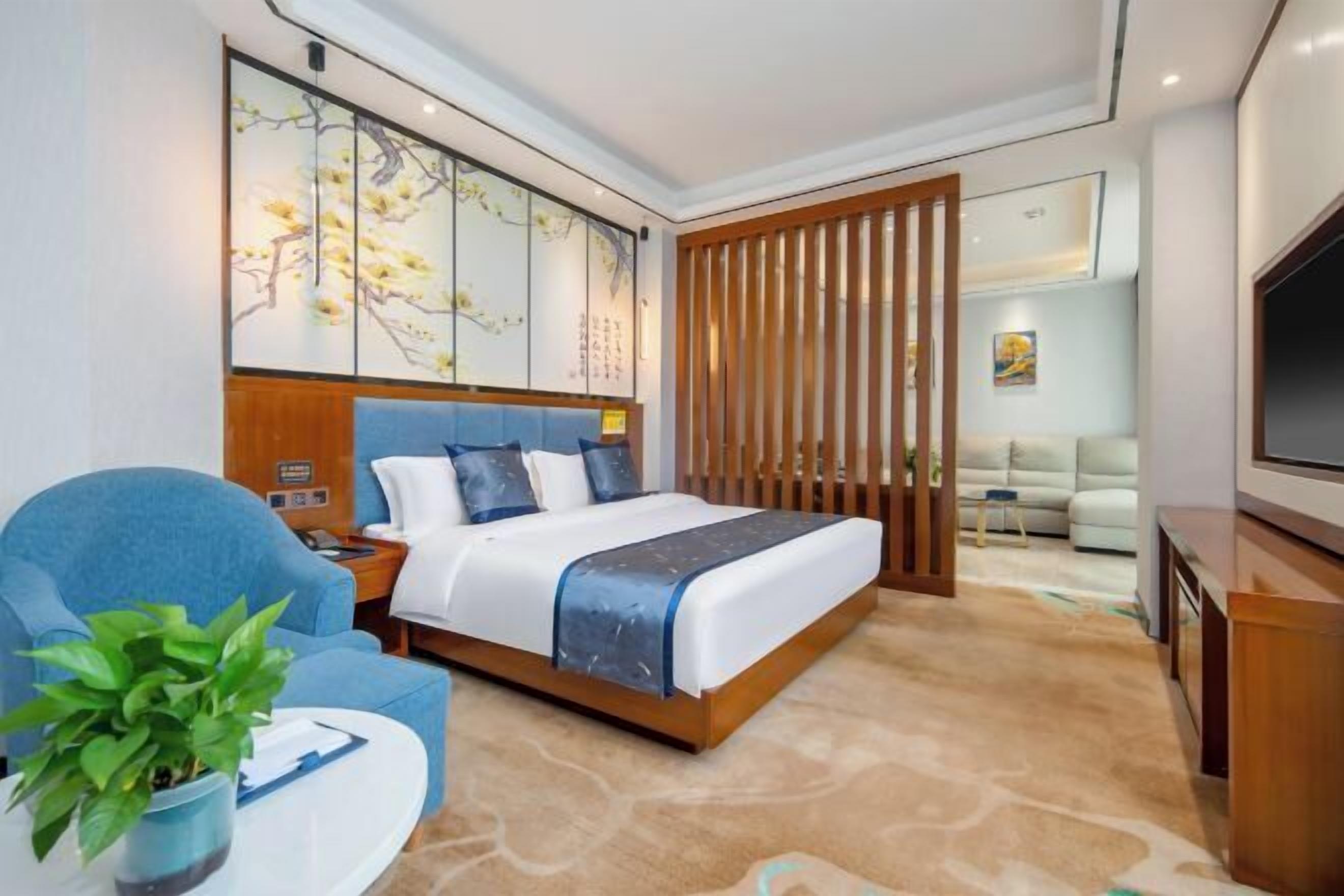 Una camera d'albergo con un letto, una poltrona blu, un tavolino con una pianta, una TV e una porta scorrevole che dà su un soggiorno.