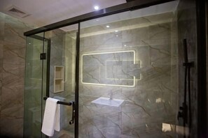 Una ducha con cerramiento de vidrio, pared de mármol, un perchero y un accesorio de iluminación geométrico.