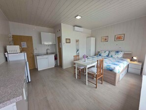 A compact room with a kitchenette, a bed with floral bedding, a table with chairs, and a bedside table with a lamp.