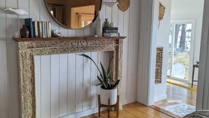 A room with a wooden floor, a fireplace with a decorative mantel, a round mirror, and a potted plant. - Dreamy Cottage right on the Lake, Dog Friendly! (Lake Waccamaw)