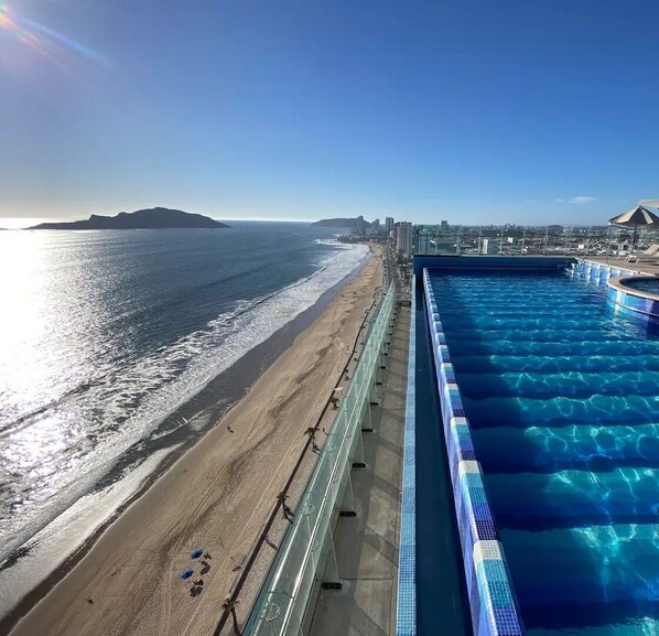 Una piscina junto a una playa, con vista al océano y montañas a lo lejos.
