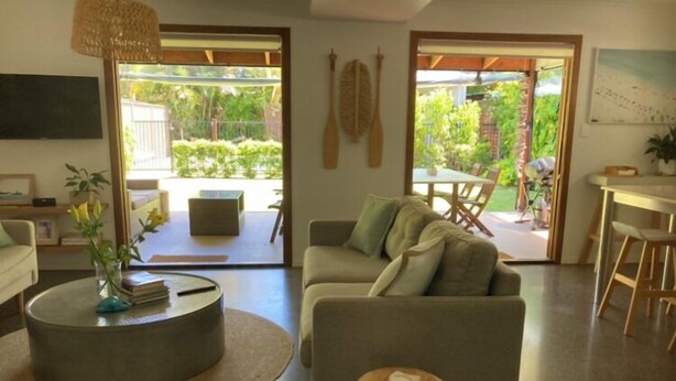 Una sala moderna con un sofá, una mesa de centro y un taburete pequeño. Se ve un comedor con una mesa y sillas a través de las puertas de cristal.