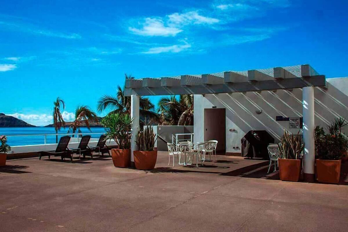 A poolside area with lounge chairs and tables, a covered seating area, and a view of the sea.