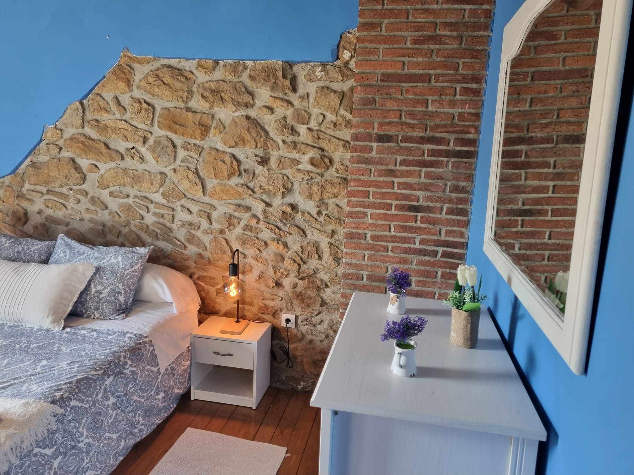 Un dormitorio con pared de piedra, pared de ladrillo, cama con ropa de cama estampada, mesita de noche con lámpara y espejo en la pared.
