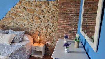 Un dormitorio con pared de piedra, pared de ladrillo, cama con ropa de cama estampada, mesita de noche con lámpara y espejo en la pared.