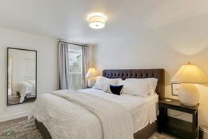 A neatly made bed with white linens and a dark headboard, two bedside lamps, a large mirror, and a window with curtains.