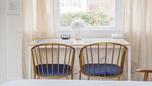 Een kamer met een raam, twee stoelen met blauwe kussens, een witte vaas met bloemen en een bureau met toetsenbord en muis.