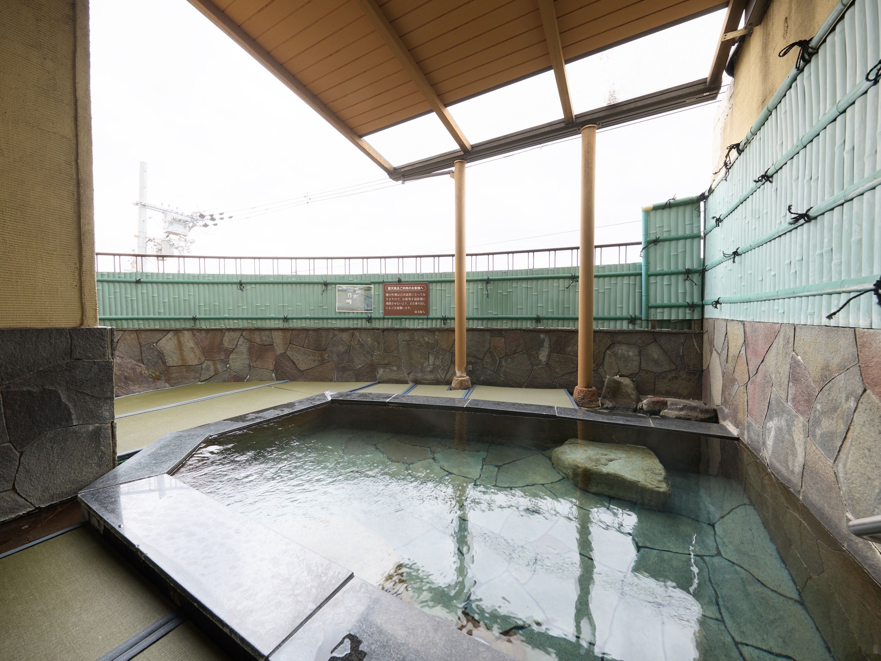 A stone-walled hot spring area with a tiled floor, a wooden roof, and a green fence in the background.