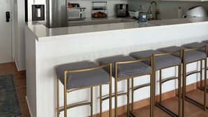 Une cuisine moderne avec des meubles blancs, un réfrigérateur en acier inoxydable, une machine à café et un évier. On y trouve quatre tabourets de bar avec des assises rembourrées.