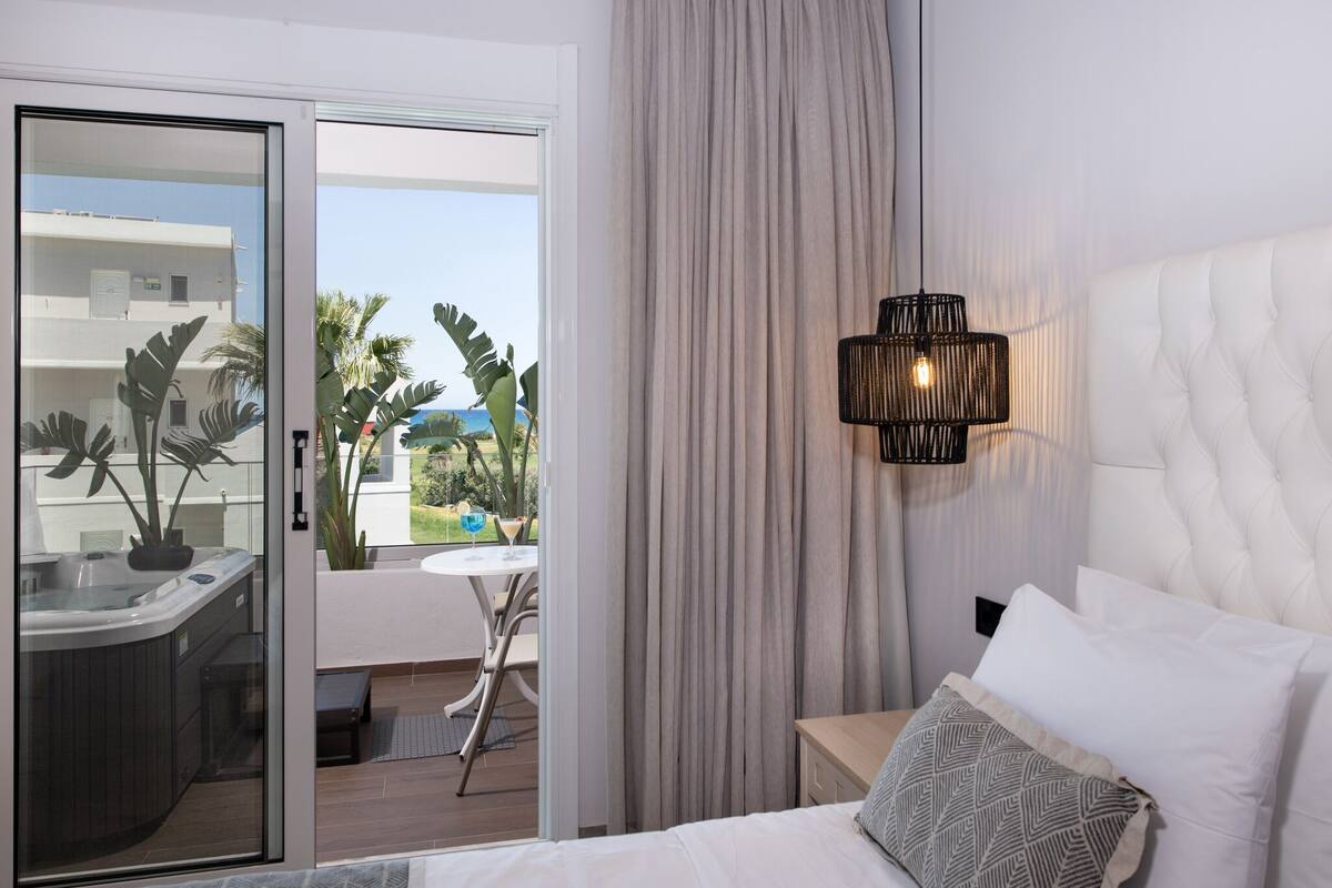 A hotel room with a bed, a nightstand, a lamp, a view of the ocean, and a balcony with a hot tub and plants.