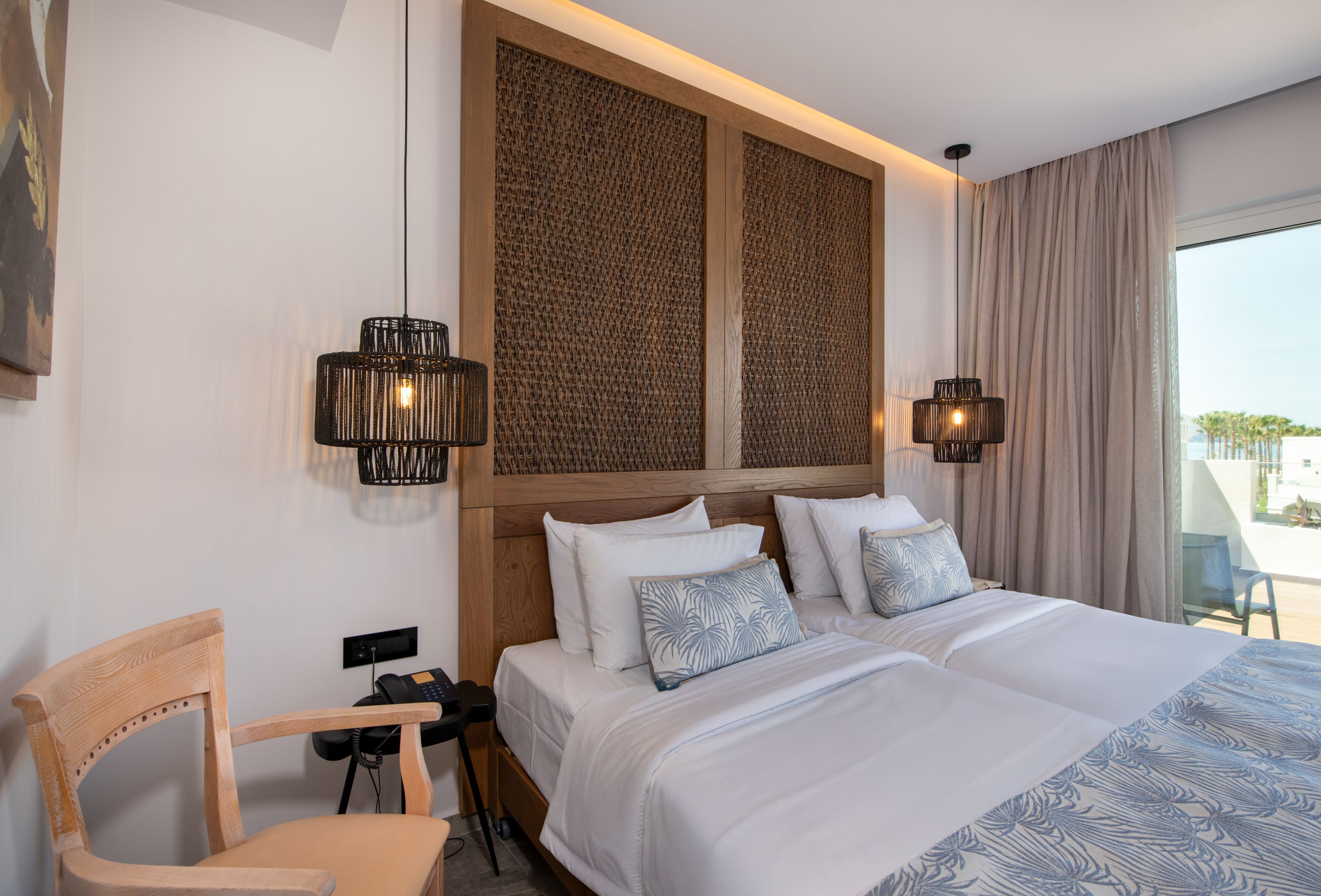 A hotel room with a bed, two bedside lamps, a chair, a desk with a phone, and a view of the outdoors.