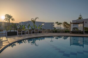 A swimming pool with lounge chairs and a pergola, surrounded by palm trees and modern buildings. - Delfina Tropic Beach (Apokoronas)