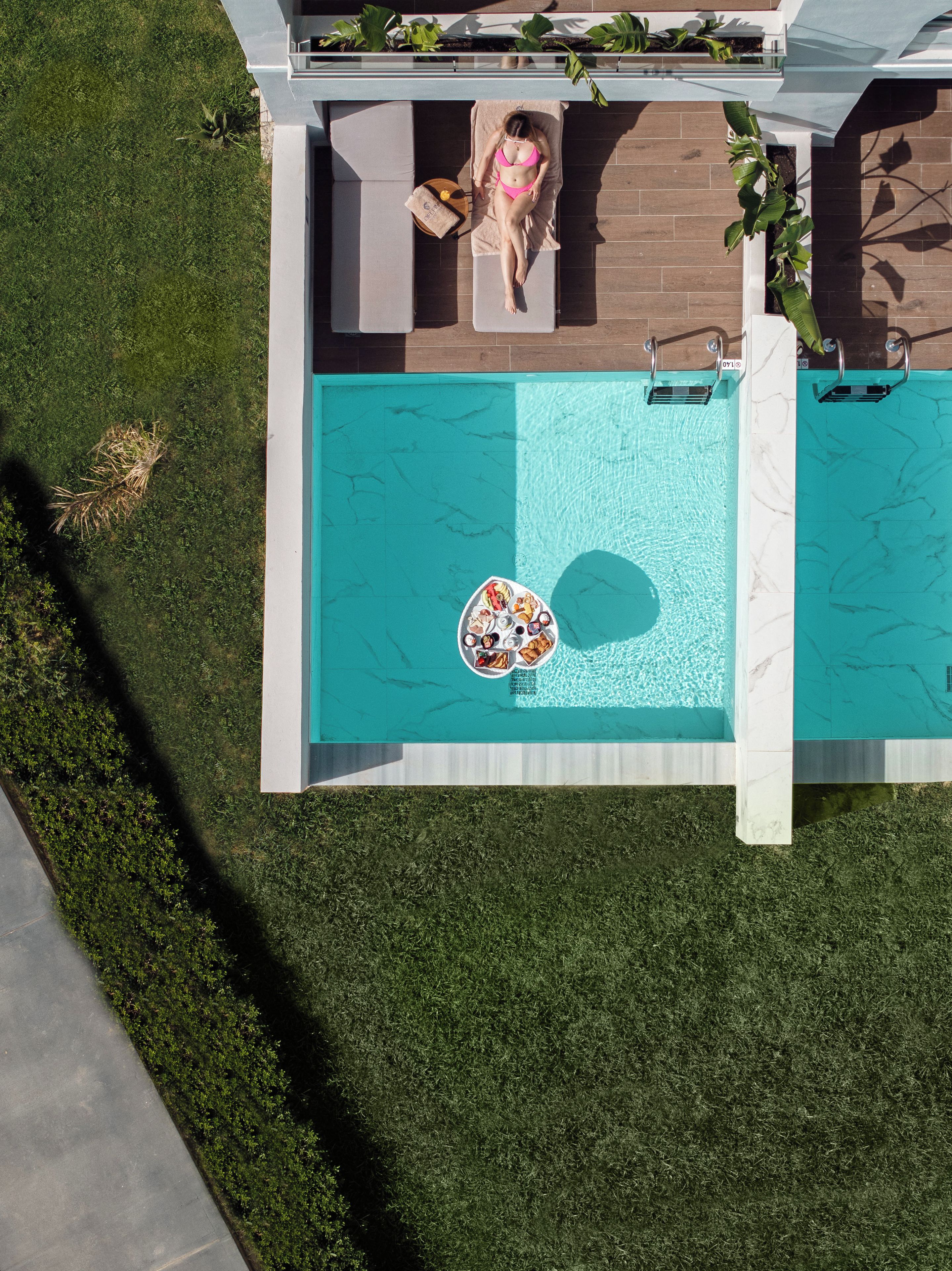 Vista aérea de una zona de piscina moderna con una silla de descanso, una mesa de billar flotante y una pequeña mesa con comida y bebidas.