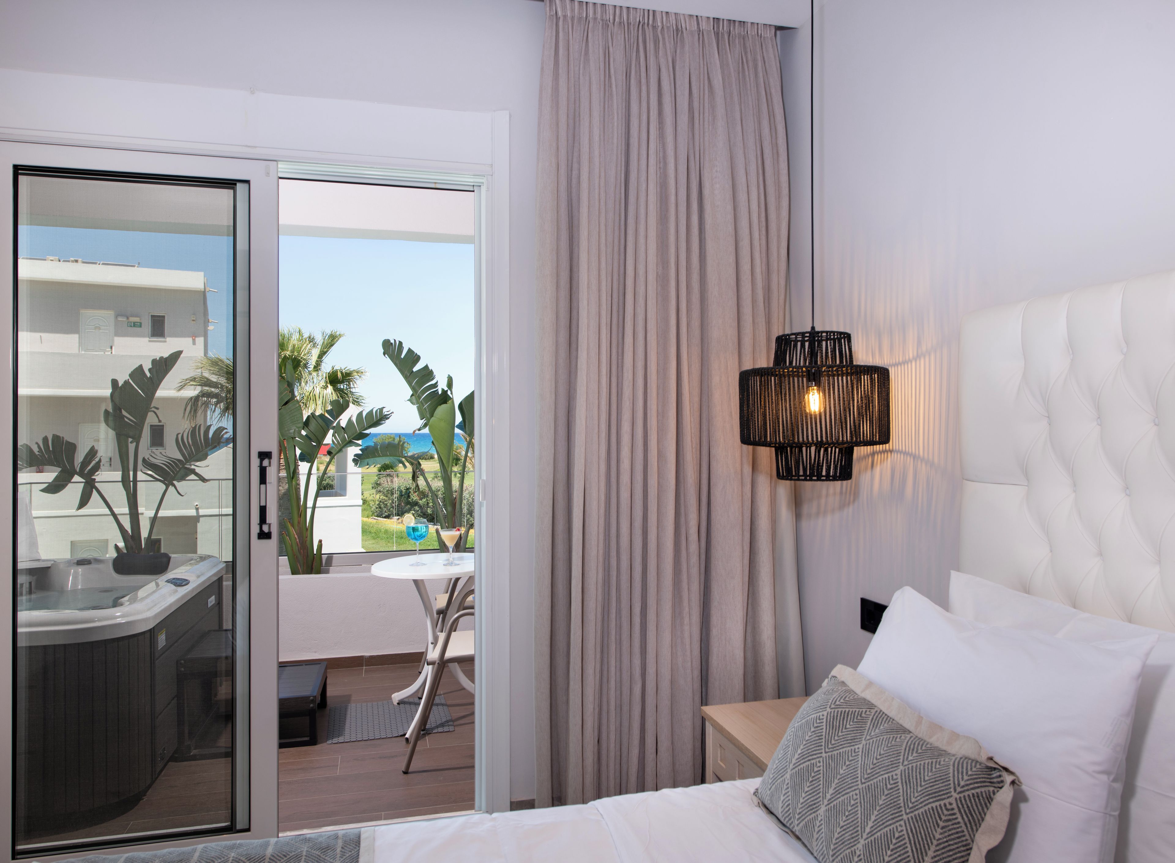 Ein Hotelzimmer mit einem Bett, einem Nachttisch, einer Lampe, Meerblick und einem Balkon mit Whirlpool und Pflanzen.