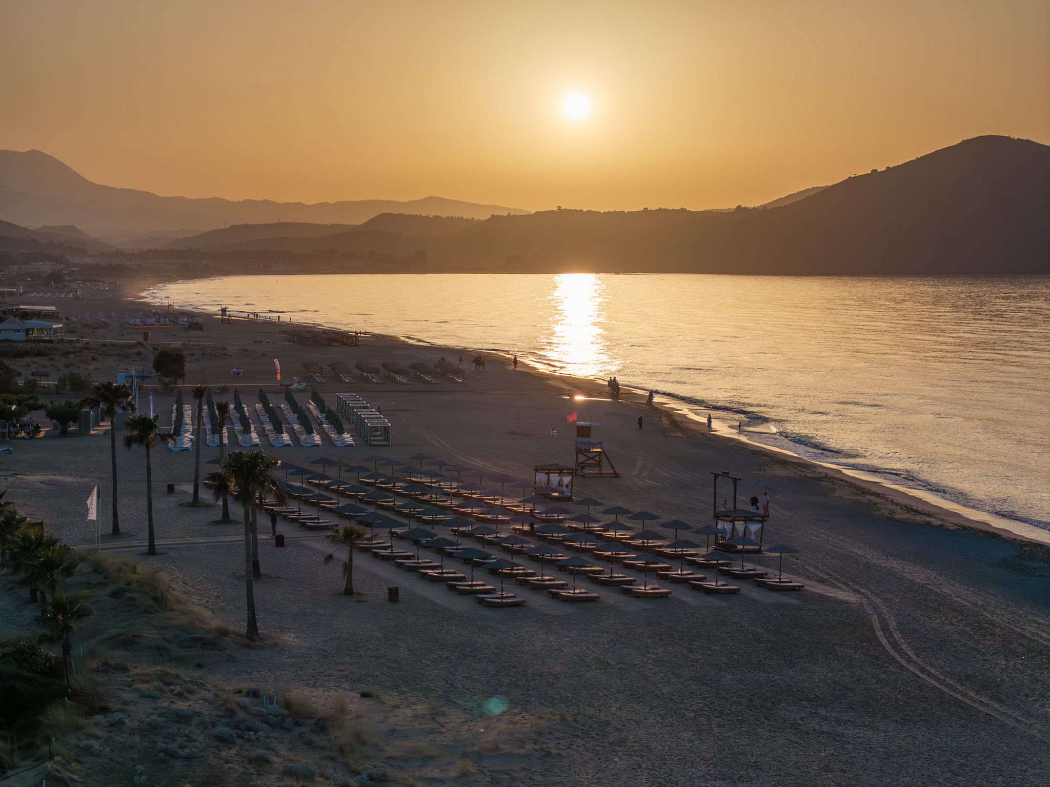 Ein Strand mit Liegestühlen und Sonnenschirmen, einem Rettungsturm und einem Sonnenuntergang über dem Wasser.