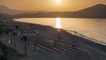 A beach with rows of sun loungers and umbrellas, a lifeguard tower, and a sunset over the water.